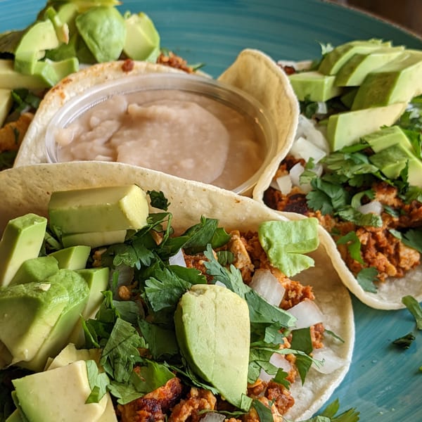 Three tacos filled with meat, cilantro, onions, and avocado, with a side of refried beans.