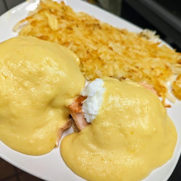 Eggs Benedict with salmon and hollandaise sauce, served with crispy hash browns on a white plate.