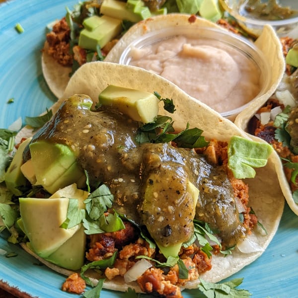 Close-up of two tacos filled with meat, avocado, cilantro, and green salsa, with a side of refried beans.