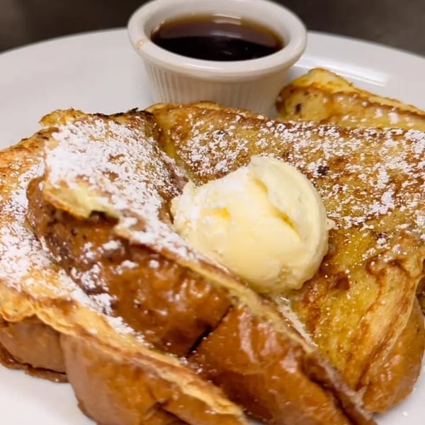 Golden French toast with powdered sugar, a dollop of butter, and a side of syrup.