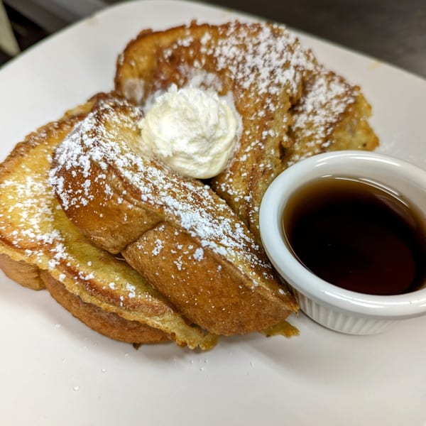 Golden French toast slices, dusted with powdered sugar, topped with whipped cream, and a side of syrup.